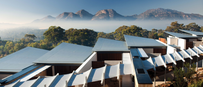 A modern architectural structure composed of multiple buildings with flat, angular roofs, connected by white awnings or canopies. In the background, a mist-covered mountain range rises behind lush green trees.