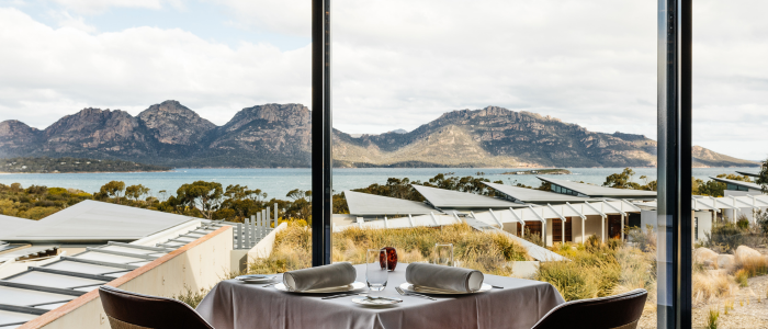 A dining table set for two, positioned by large windows overlooking a scenic landscape with mountains, water, and modern buildings with flat roofs. The table includes two chairs, white napkins, plates, cutlery, and a small decorative centerpiece.