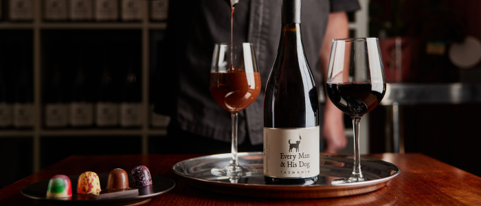 A bottle of wine labeled 'Every Man & His Dog Tasmania' placed on a silver tray along with two glasses of wine. One glass is being filled with what appears to be chocolate. In the foreground, there is a plate with four colorful chocolates. The background includes shelves with bottles and a person standing behind the tray.