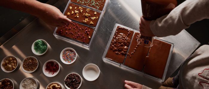 Two people making chocolate bars. One person is pouring melted chocolate into molds, while the other is adding various toppings to the chocolate. There are several small bowls containing different toppings such as green sprinkles, rainbow sprinkles, nuts, dried fruits, and chocolate chips on the table.
