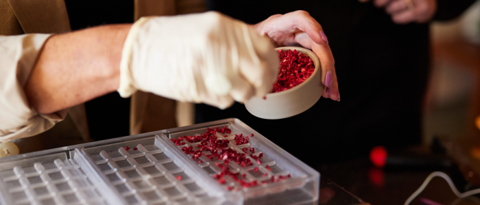 A person wearing a glove and holding a small bowl filled with red pieces, possibly some kind of food ingredient or material. The person is pouring these red pieces into a mold or tray with multiple compartments.