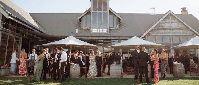 A group of formally dressed people gathered on a grassy lawn outside a modern building with large windows and a peaked roof. White canopy tents provide shade as guests mingle. Wooden barrels and outdoor furniture are scattered around the area.