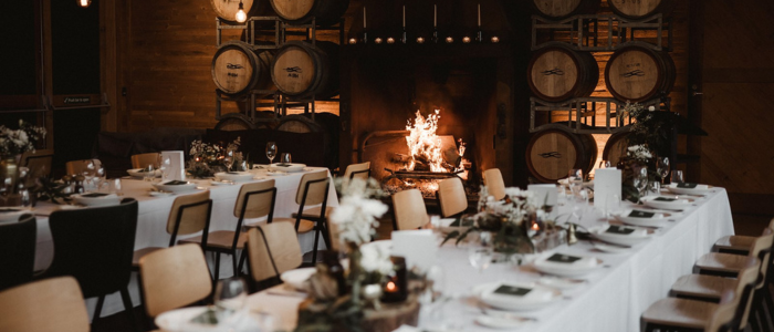 A warmly lit dining area featuring long tables draped in white tablecloths, adorned with floral centerpieces and candles. Wooden chairs surround the tables. A large fireplace with a burning fire adds a cozy ambiance, and stacked wine barrels behind it enhance the rustic charm.
