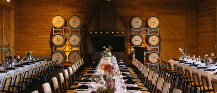 A rustic indoor dining setup featuring two long, parallel tables with place settings including plates, glasses, and napkins. Floral arrangements decorate the tables. In the background, a fireplace is flanked by stacked wine barrels on metal racks, enhancing the cozy, winery-inspired ambiance.