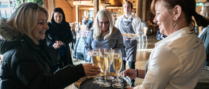 An indoor social gathering in a warmly lit wooden interior. A server holds a tray of champagne or sparkling wine as a guest reaches for a glass. Another server in the background carries a tray of appetizers. The scene suggests a reception or celebratory event, with several attendees present.