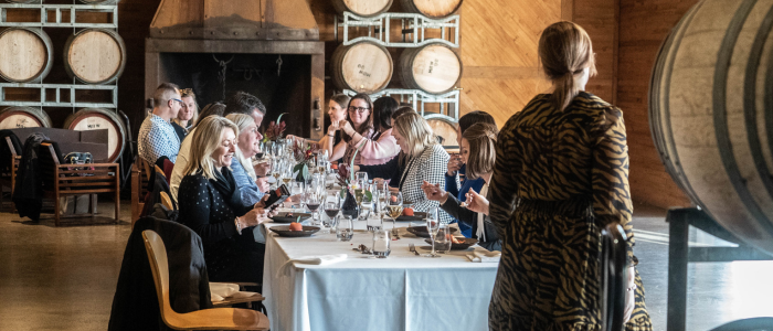 A group of people seated around a long dining table in a winery or wine cellar, surrounded by stacked wine barrels on racks. The table is set with plates, glasses, and cutlery, and the guests appear to be enjoying a meal and conversation.