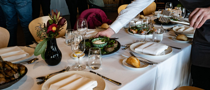 A dining table set for a meal with a white tablecloth, place settings including plates, napkins, cutlery, and glasses. Various dishes, including a bowl of salad, are arranged on the table. A person is reaching over to adjust or place something. A black vase with flowers sits on the left, and people are visible in the background around another table.