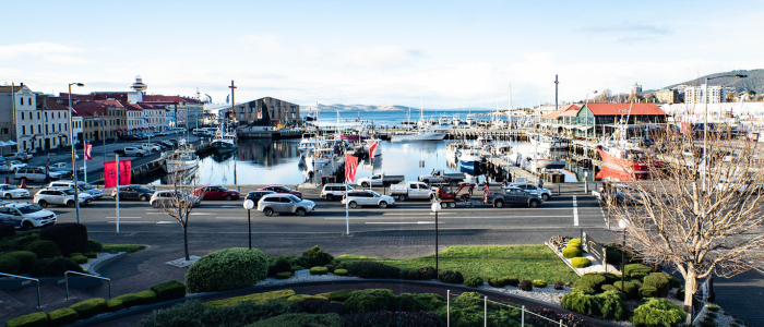 A scenic view of a harbor with several boats docked in the water. Buildings line both sides of the harbor, with a street featuring parked cars and light traffic in front. The sky is mostly clear with some clouds, and mountains are visible in the background. The foreground includes landscaped greenery and a few bare trees.