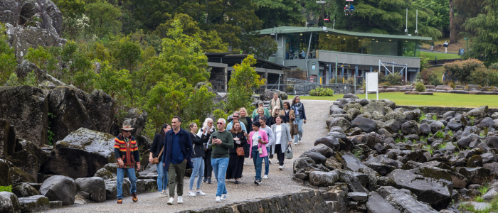 A group of people walking on a path surrounded by rocks and greenery. In the background, there is a building with large windows and a green roof, possibly an entrance or visitor center. The setting being the Launceston Gorge.