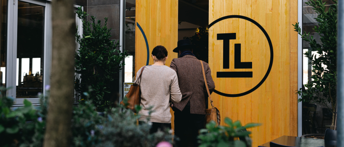 Two people walking into a building with a large wooden door featuring a circular logo with the letters 'TL' in the center. Plants flank the entrance, and the setting appears to be urban.