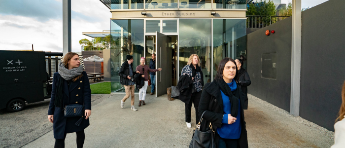A group of people walking out of a building with glass doors labeled 'Ether Building.' A black truck on the left side displays the text 'X+ Museum of Old and New Art.