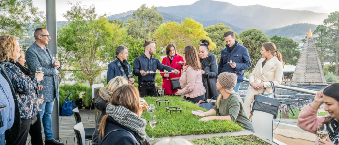 A group of people gathered outdoors on a terrace or patio, surrounded by greenery and distant mountains. They are standing and sitting around a table with grass on it, conversing and holding drinks. Some are wearing coats and jackets, indicating cool weather.