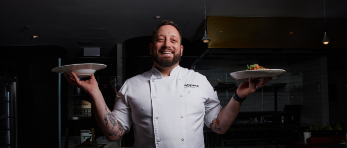 A person wearing a white chef's jacket standing in a kitchen. The person is holding two plates, one in each hand. There are various kitchen appliances and utensils visible in the background.