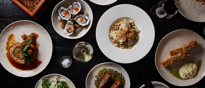 A variety of plated dishes arranged on a dark table. There are several plates with different types of food, including oysters with orange garnish, a pasta dish topped with cheese and breadcrumbs, a rolled meat dish with sauce and greens, two pieces of bread with a green sauce and white spread, and other assorted dishes. There are also glasses of white wine and red wine on the table.