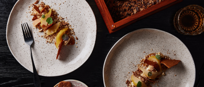 Two plates of a dessert arranged on a dark wooden table. Each plate contains a portion of the dessert, which appears to be composed of thin slices of fruit, possibly apple or pear, layered with what looks like caramelized or baked elements and garnished with mint leaves. There is also a fork placed on the left plate. In the background, there is an open wooden box filled with what seems to be chocolate shavings or crumbles and a glass containing an amber-colored liquid, likely whiskey or another type of drink.