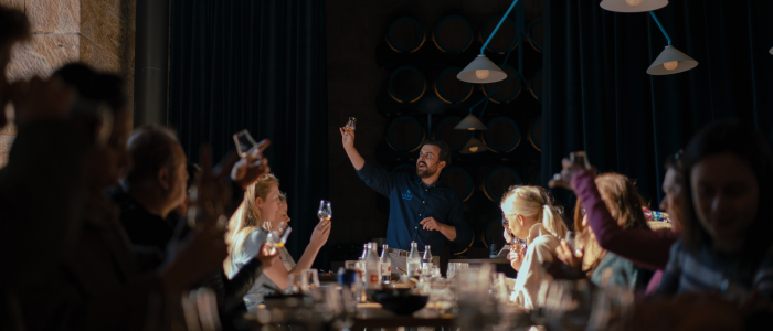 A group of people sitting around a table, raising their glasses inspecting their whiskey. The setting appears to be indoors, possibly in a restaurant or bar with dim lighting. Bottles and glasses are on the table, and barrels are stacked against the wall in the background. One person stands at the center, leading the demonstration and teaching those at the table about the whiskey