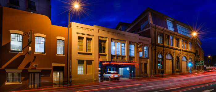 A nighttime street scene with three buildings. The building on the left is brown with arched windows and a door. The middle building is beige with rectangular windows. A car is parked under an illuminated red sign at the entrance of this building. The building on the right is larger, with more intricate architectural details and large arched windows. Streetlights illuminate the scene, and light trails from passing vehicles are visible on the road in front of the buildings.