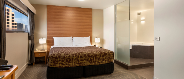 A modern hotel room with a neatly made bed featuring white pillows and a brown patterned bedspread. There are two bedside tables, each with a lamp. The room has a large window with curtains on the left side, and there is an open bathroom area with glass walls on the right side, revealing a bathtub. The overall decor is contemporary and clean.
