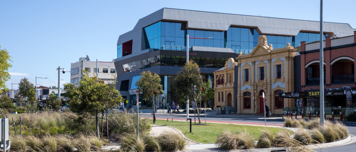 A modern urban area featuring a landscaped foreground with grass and small trees. The background includes a mix of contemporary and historical buildings, such as a large glass-fronted structure with angular design and an older building with classical columns and ornate details.