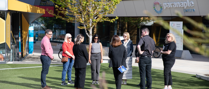 A group of people standing in a circle on a grassy area outside a building with signage reading 'paranaple centre'. The group are engaged in conversation