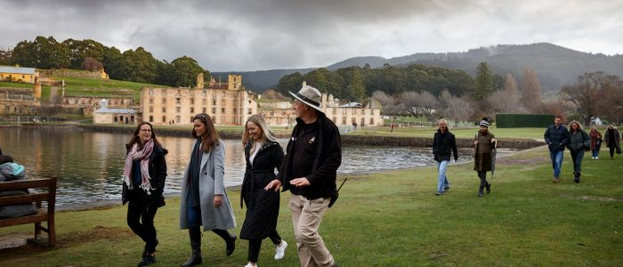 A group of people walking along a grassy area near a body of water with historic buildings in the background. The sky is overcast, and there are hills and trees in the distance. Some people are wearing coats and hats, suggesting cool weather. The location is Port Arthur Historical Site