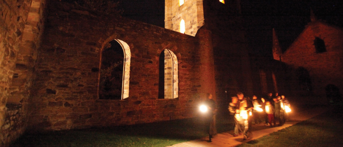 A group of people walking at night along the exterior of an old stone building with arched windows. The people are holding lanterns, illuminating their path as they walk on a paved walkway next to the building.