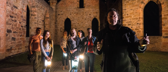 A group of people standing inside what appears to be the ruins of an old stone building at night. The people are holding lanterns and flashlights, illuminating their surroundings. The building has tall, arched windows and walls made of brick and stone.