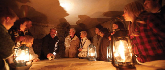 A group of people gathered around a table in a dimly lit room. There are several lanterns on the table providing light. The room appears to be made of stone, suggesting it might be an old or historical setting such as a cellar or crypt.