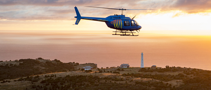 A blue helicopter flies over a coastal landscape at sunset, with a lighthouse and several buildings nestled among vegetation below. The ocean stretches into the background, illuminated by the warm glow of the setting sun.