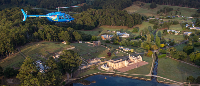 A blue helicopter flies over Port Arthur Historical Site featuring multiple buildings, open fields, and wooded areas. The estate is surrounded by greenery, with a canal running through the property and a body of water in the foreground.