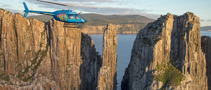 A blue helicopter flies between two tall, rugged rock formations with a body of water in the background. The steep cliffs frame a distant landscape of hills or mountains.