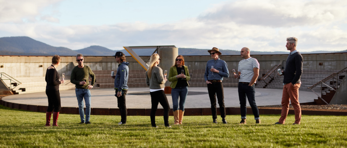 A group of eight casually dressed people standing on a grassy area, conversing near a large circular concrete structure. The background features partly cloudy skies and distant mountains.