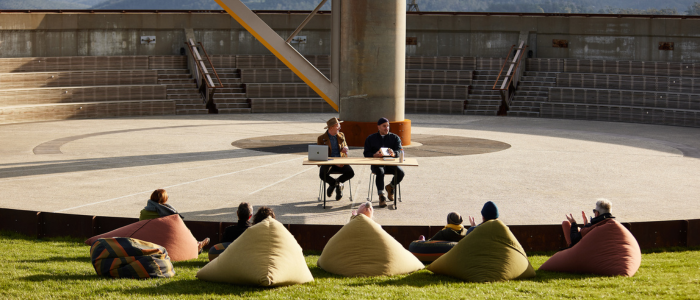 An outdoor amphitheater with a circular stage where two people sit at a table with laptops. Several others are seated on bean bags on the grass, facing the stage. The background features tiered seating and a large structural column at the center of the stage area.