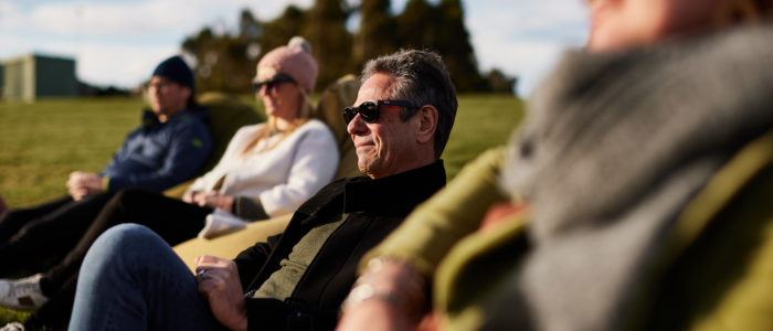 Four people sitting on a grassy area outdoors, dressed casually. The background features trees and a possible building or structure on the left.