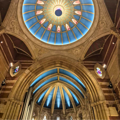 Interior of St John’s Church in Launceston, highlighting a grand dome ceiling with blue and gold patterns, surrounded by large stone arches with blue accents. Stained glass windows flank the arches, and the architecture features Gothic elements with intricate carvings and vibrant colors.