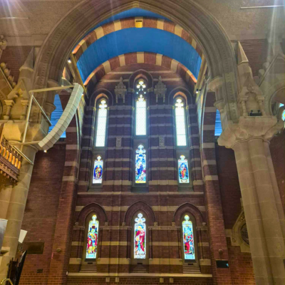 Interior of St John’s Church in Launceston, featuring tall arched stained glass windows with vibrant depictions of religious figures and scenes. The architecture includes striped brick walls, ornate columns, and a blue ceiling above the windows.