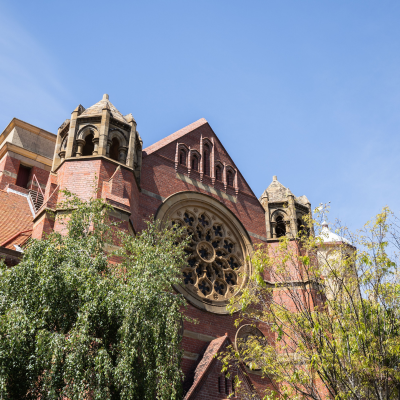 St Johns Church - a red brick building with Gothic architectural features, including two pointed towers and a large circular stained glass window. Trees partially obscure the building in the foreground, and the sky is clear and blue.
