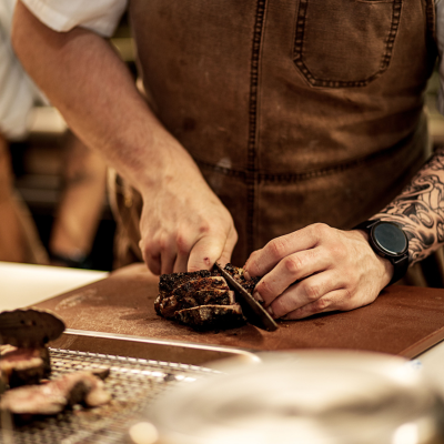 A person wearing a brown apron and smartwatch slices a piece of seared meat on a wooden cutting board, with a tattooed arm visible and cooked meat pieces resting nearby on a metal rack.