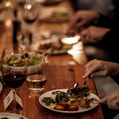 A close-up of a dinner table with several plates of food and glasses of wine. The focus is on a plate with roasted vegetables, greens, and meat, along with a glass of red wine and a glass of water. Other diners' hands are visible in the background, holding utensils and eating. The table is wooden, and there are small name cards placed near the plates.