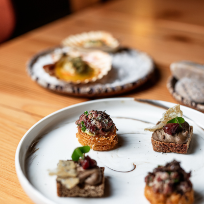 A close-up of a plate with various gourmet appetizers. The plate in the foreground contains four different bite-sized pieces, each garnished with herbs and other toppings. In the background, there are additional plates with more appetizers, including what appears to be scallops served on their shells and another dish with bread or pastries. The setting is on a wooden table.