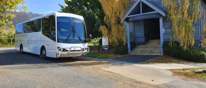 A white bus parked on the side of a road near a building with a sign reading 'Frogmore Creek.' The building has steps leading to the entrance, surrounded by trees with yellow leaves.