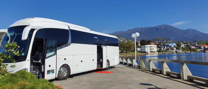 A large white tour bus with its door open, parked on a concrete surface near a body of water. Mountains rise in the background, with buildings and houses along the waterfront under a clear blue sky. Plants are visible in the foreground.