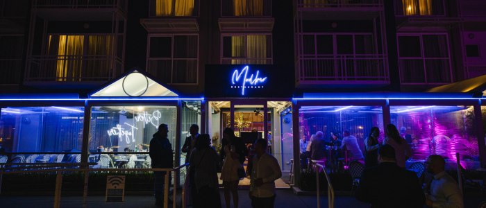 Nighttime view of the exterior of 'Mud Bar Restaurant,' featuring modern architecture with blue neon lighting. People are gathered outside, while others are visible inside under purple and blue lighting.