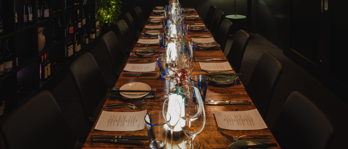 A long dining table set for a formal meal, featuring plates, glasses, cutlery, and menus at each seat. Wine bottles are displayed on shelves along the left wall, and the dim lighting creates an intimate ambiance.