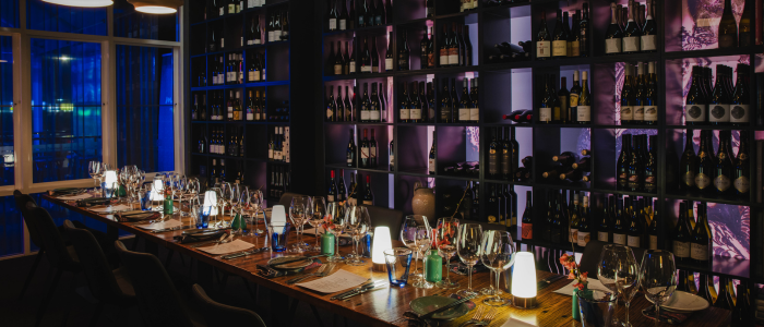 A long dining table set for a meal in a wine-themed restaurant or cellar, featuring wine glasses, plates, cutlery, and small lamps. Shelves filled with wine bottles line the background, and the dim lighting creates an elegant, intimate ambiance.