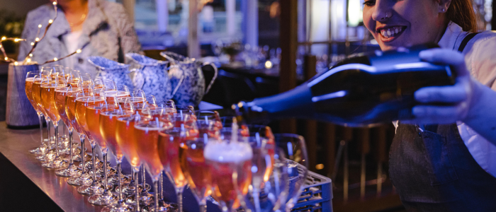 A person pouring champagne into a row of glasses on a bar counter in a dimly lit setting, with fairy lights and ceramic pitchers in the background, suggesting a festive or celebratory atmosphere.