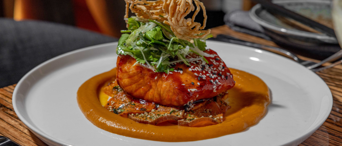 A plated dish featuring glazed salmon topped with sesame seeds, greens, and crispy fried noodles, served on a bed of thinly sliced vegetables with an orange-colored sauce, all presented on a white plate.