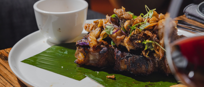 A plated dish featuring grilled meat topped with sautéed vegetables and microgreens, served on a green leaf with a small white cup on the side. A glass of red liquid, possibly wine, is partially visible in the foreground.