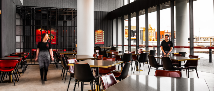 Interior of a modern restaurant or café with large windows letting in natural light. Several tables and chairs are neatly arranged, with two staff members in black uniforms—one walking and the other cleaning or arranging a table. The décor features red and black elements, a large white pillar in the center, and a wall sign displaying the repeated text "Goodguy".