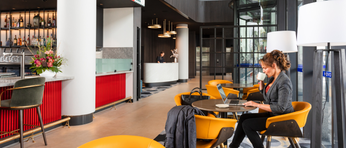 Modern, stylish interior of Mr. Goodguy in Devonport. A bar with shelves of bottles and glasses is on the left, with a person working behind the counter. In the foreground, several round tables with bright yellow chairs are arranged, and a person is seated at one table using a laptop. The background includes large windows, tall columns, and additional seating areas.